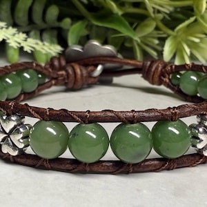 May include: A handcrafted bracelet featuring green jade beads and brown leather. The bracelet has a wrap-around design with silver-colored clover charms. The closure is a metal clasp. The jewelry is suitable for everyday wear.