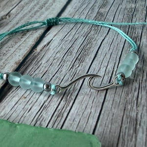 May include: A light blue cord bracelet featuring a central silver-toned metal wave charm. The design includes three frosted sea glass-style beads on each side of the wave, separated by small silver-toned metal spacer beads along the cord.