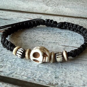 May include: A black macrame cord bracelet featuring a central off-white skull bead flanked by two smaller patterned beads and four smooth round spacer beads. The bracelet has adjustable black cord ends for a secure fit around the wrist.