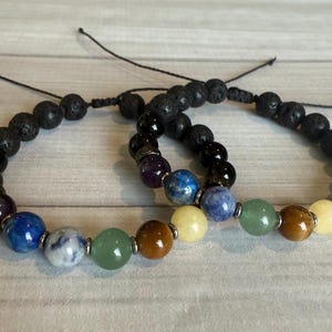 May include: Two beaded bracelets featuring a chakra design. The bracelets have black lava rock beads and colorful gemstone beads in a rainbow pattern. The bracelets are strung on a black cord.