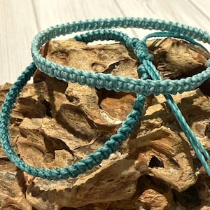 May include: Two turquoise macrame bracelets are displayed on a textured, light brown surface. The bracelets are made of a tightly woven cord, with a knotted design. The bracelets are a light blue-green color, and the background is a natural wood.