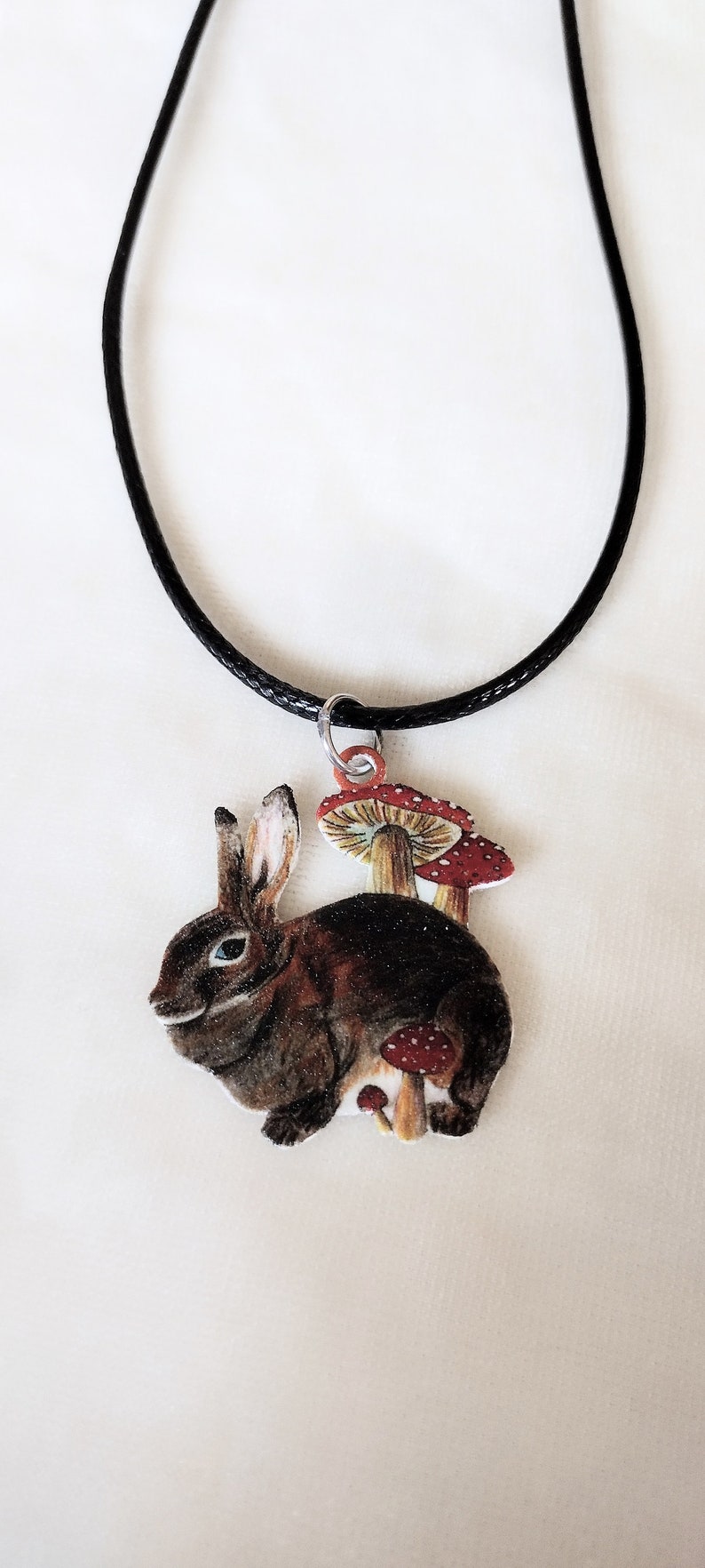 May include: A black cord necklace with a brown rabbit charm. The rabbit is surrounded by red and white mushrooms.