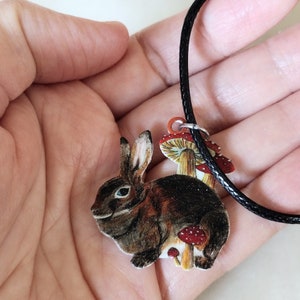 May include: A brown rabbit with a white belly and blue eyes sits on a mushroom with red and white spots. The rabbit is part of a necklace with a black cord.