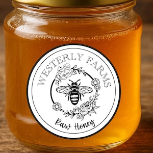 May include: A small glass jar filled with golden raw honey. The jar has a gold lid and a white label with the text "Westerly Farms Raw Honey" and a bee illustration. The jar is set on a wooden surface.
