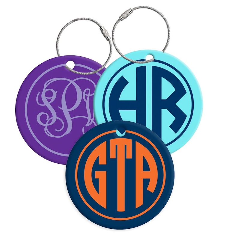 May include: Three personalized luggage tags with silver-tone wire loops. One is purple with a white monogram, another is light blue with dark blue initials, and the third is navy blue with orange initials.
