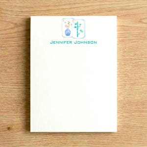 May include: A white rectangular notepad featuring a blue and white floral vase illustration next to a green bamboo stalk illustration at the top center. The name JENNIFER JOHNSON is printed in teal capital letters below the illustrations.
