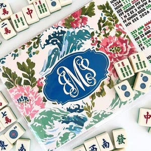 May include: A collection of ivory-colored Mahjong tiles scattered around a floral-patterned case. The case features a blue oval with the initials "MS" and a pink, green, and blue floral design. The tiles have Chinese characters and symbols.