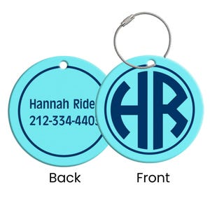 May include: A light blue luggage tag with a silver-colored wire loop. The front features a dark blue monogram within a circle. The back has the name "Hannah Ride" and a phone number.