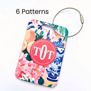 May include: A rectangular luggage tag with a floral design featuring oranges, pink flowers, and blue vases. A red circle with the letters "TOT" is in the center. The tag has a silver wire loop for attachment. The text "6 Patterns" is at the top.