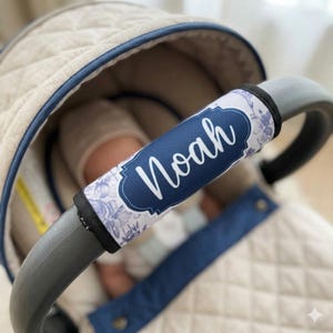 May include: A custom stroller handle cover featuring a white and blue floral pattern with a dark blue central label displaying the name Noah in white script. The cover is secured around the curved grey handle of a quilted beige baby carrier.
