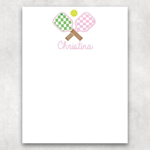 May include: A white stationery sheet featuring a graphic of two crossed pickleball paddles with brown handles, one green checkered and one pink checkered, with a yellow pickleball above them and the name Christina written in pink script below.