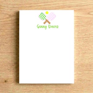 May include: A white rectangular notepad featuring a graphic of two crossed pickleball paddles in green and pink checkered patterns with brown handles, a small yellow pickleball, and the name Ginny Davis printed in green script font below.