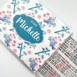 May include: A personalized planner with a teal label that reads "Michelle" and the year "2026". The cover features a white background with a pattern of blue birds, pink flowers, and bamboo stalks. The planner is open, revealing a section with contact information.