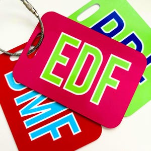 May include: Three rectangular luggage tags with rounded corners and metal cable loops. The top tag is hot pink with lime green letters reading EDF. The bottom left tag is red with light blue letters. The top right tag is lime green with blue letters.