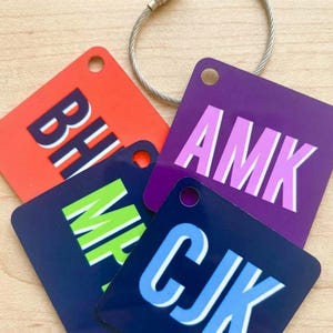 May include: Four square acrylic luggage tags with rounded corners and a metal cable loop. The tags feature bold, block-letter monograms: orange with BH, purple with AMK, dark blue with MP, and dark blue with CJK, each in contrasting colors.