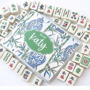 May include: A personalized 2026 planner with a green and white floral design, featuring the name "Katy." The planner is surrounded by ivory Mahjong tiles with red, green, and black symbols.