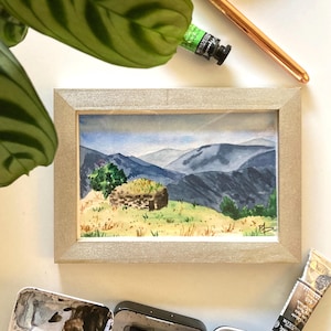 May include: A watercolor painting of a small stone house with a thatched roof, nestled in a green valley with mountains in the background. The painting is framed in a silver frame.