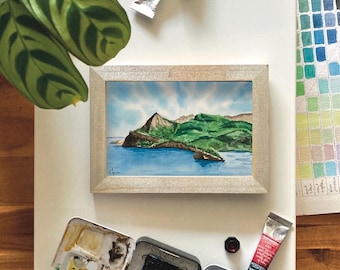 May include: A framed watercolor painting of a green island with a rocky coastline and blue water. The painting is on a white surface with a variety of art supplies, including paint tubes, a watercolor palette, and a paintbrush.