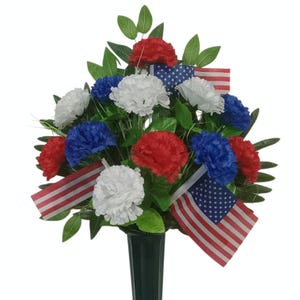 May include: A floral arrangement featuring red, white, and blue carnations interspersed with green leaves and miniature American flags. The flowers are set in a black, tapered, conical vase, creating a patriotic display of artificial blooms.