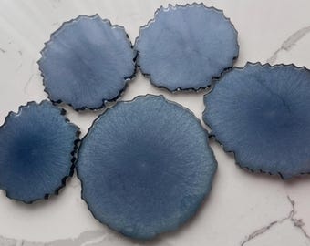 May include: Five blue resin coasters with a unique, organic shape. Each coaster has a dark blue, textured edge and a lighter blue interior, creating a marbled effect. These decorative coasters are designed to protect surfaces from drink rings.