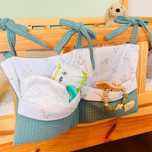 Op de afbeelding: Een turquoise en witte hangende organizer met drie vakken. De organizer hangt aan een houten wieg. De vakken bevatten babydoekjes, een luier en een houten bijtring met kralen.