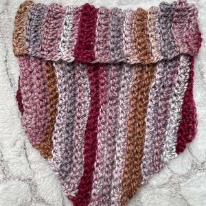 May include: A handmade, chunky knit cowl in a triangle shape. The cowl features vertical stripes of varying colors, including shades of pink, burgundy, brown, and gray. The thick yarn creates a textured appearance, perfect for cold weather.