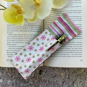 May include: A fabric bookmark with a pen holder, featuring a floral pattern with pink flowers on a white background. The top section has pink and green stripes. The bookmark is open on a book with yellow flowers.