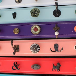 May include: Five wooden shelves painted in different colors, each with a different decorative knob and hook. The shelves are painted white, light blue, purple, pink, and red. The knobs are a variety of styles and colors, including a heart, a flower, a clock, a swirl, and a dog. The hooks are all black metal and are in different shapes, including a single hook, a double hook, and a triple hook.