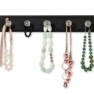 May include: A black rectangular wall-mounted rack featuring five clear crystal-style knobs. Hanging from the knobs are various necklaces, including a pearl strand with a beige ribbon, a black beaded chain, a pale green bead necklace, a pink ringed necklace, and a green bead necklace.