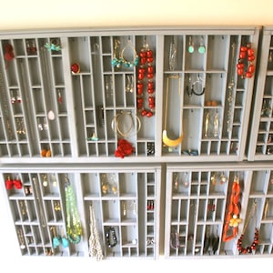 May include: A wall-mounted gray wooden printer drawer organizer featuring numerous small compartments used to display various jewelry items including necklaces, earrings, bracelets, and rings hanging from small metal hooks within the grid.