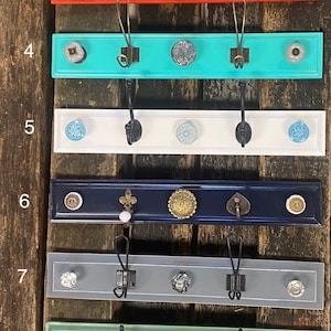 May include: A collection of ten decorative coat racks, each featuring a painted wooden board with three hooks and unique decorative knobs. Colors include teal, green, red, white, navy, and gray. The racks are mounted on a rustic wooden background.