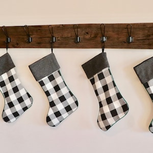 May include: Four black and white buffalo plaid Christmas stockings with gray cuffs hanging on a rustic wooden rack with metal hooks.