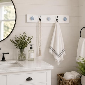 May include: A white wall-mounted rack featuring four black metal hooks and three decorative blue and white patterned ceramic knobs. The rack is mounted on a white shiplap wall, holding a white towel with black stripes and a beaded tassel.