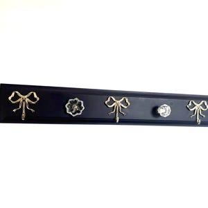 May include: A black wall-mounted coat rack with four hooks. Two gold-colored bow-shaped hooks flank a clear, faceted knob and a clear, flower-shaped knob. The rack is rectangular and mounted on a white wall.