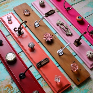 May include: A collection of decorative wall-mounted coat racks in various colors, including red, coral, pink, and natural wood. Each rack features unique knobs and hooks, showcasing a variety of styles and materials for hanging items.