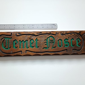 Puede incluir: Un letrero rectangular de madera con un acabado teñido en marrón oscuro y veta visible. El texto "Temet Nosce" está tallado en el centro con letras hundidas pintadas de verde. Una regla metálica muestra una longitud de 30 cm.