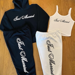 May include: A black hoodie and sweatpants set, and a white tank top and sweatpants set. All pieces have the text "Just Married" in a cursive font. The outfits are displayed on a wooden surface.