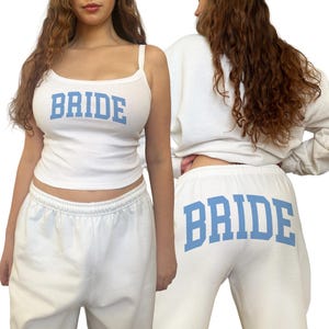 May include: White bridal loungewear set featuring a tank top and sweatpants. The tank top has thin straps and the word "BRIDE" in light blue block letters. The sweatpants also display "BRIDE" in the same font and color.
