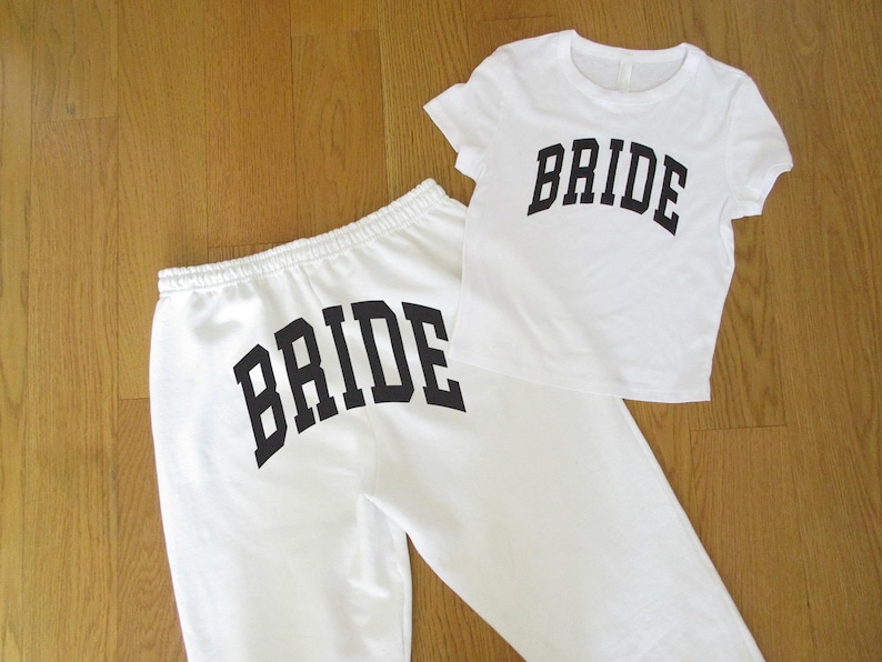 May include: White sweatpants and a white t-shirt, both with the word "BRIDE" printed in large, black, arched letters. The t-shirt is short-sleeved. The sweatpants have a drawstring waist. The items are displayed on a light brown wooden surface.