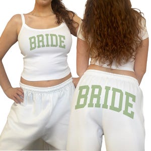 May include: White tank top and sweatpants set with the word "BRIDE" in green block letters. The tank top has spaghetti straps, and the sweatpants have an elastic waistband. The outfit is perfect for a bachelorette party or bridal shower.