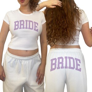 Pu&ograve; includere: Completo crop top e pantaloni della tuta bianchi. La parola "BRIDE" &egrave; stampata in lavanda sulla parte anteriore del top e sul retro dei pantaloni. Un outfit casual, perfetto per un evento nuziale.