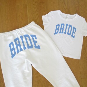 Pu&ograve; includere: Un completo composto da maglietta e pantaloni della tuta bianchi. La parola "BRIDE" &egrave; stampata in lettere blu sulla parte anteriore della maglietta e sul retro dei pantaloni della tuta. Il set &egrave; esposto su una superficie di legno marrone chiaro.