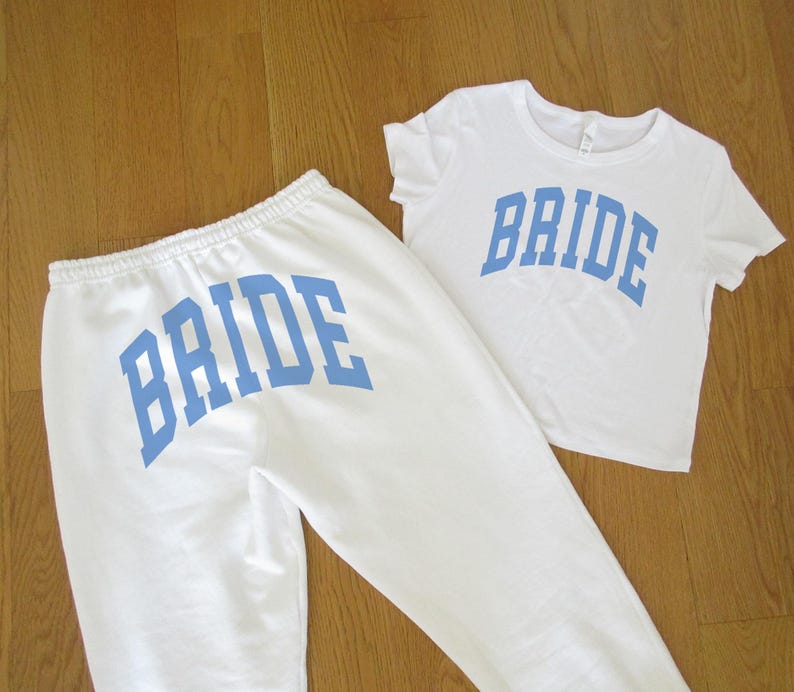 Pu&ograve; includere: Un completo composto da maglietta e pantaloni della tuta bianchi. La parola "BRIDE" &egrave; stampata in lettere blu sulla parte anteriore della maglietta e sul retro dei pantaloni della tuta. Il set &egrave; esposto su una superficie di legno marrone chiaro.