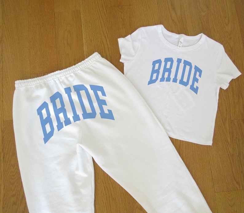 May include: A white t-shirt and sweatpants set with the word "BRIDE" in light blue block letters. The t-shirt is short-sleeved, and the sweatpants have a drawstring waist. The set is likely for a bridal event.