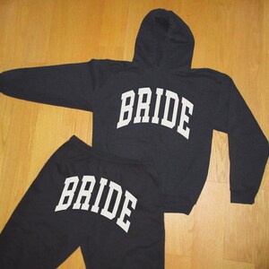 Pu&ograve; includere: Completo nero composto da felpa con cappuccio e pantaloni della tuta abbinati, entrambi con la scritta "BRIDE" in lettere bianche e in grassetto. La felpa ha un cappuccio e maniche lunghe. Il completo &egrave; presentato su una superficie di legno.