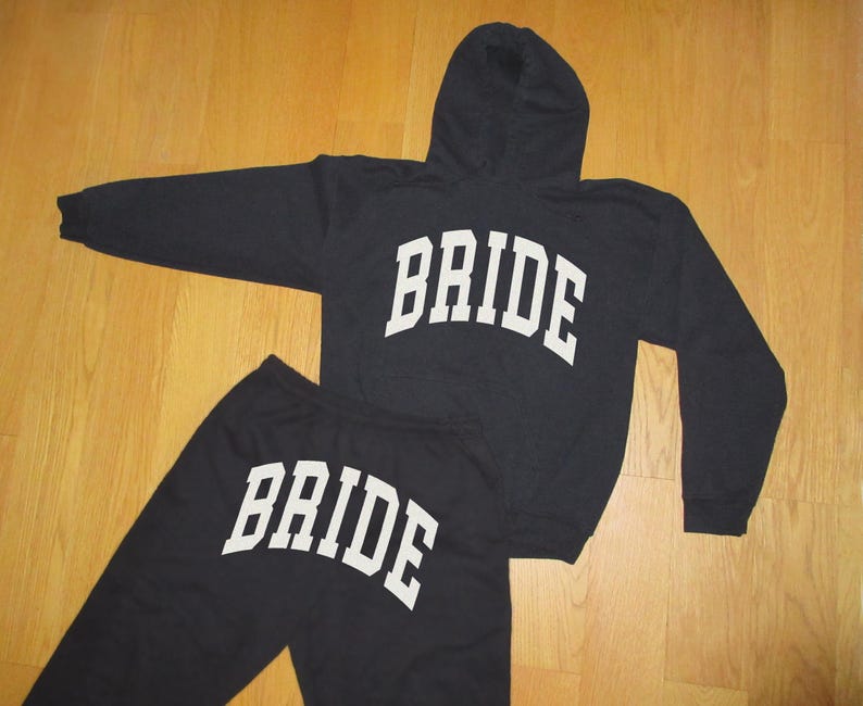 Pu&ograve; includere: Completo nero composto da felpa con cappuccio e pantaloni della tuta abbinati, entrambi con la scritta "BRIDE" in lettere bianche e in grassetto. La felpa ha un cappuccio e maniche lunghe. Il completo &egrave; presentato su una superficie di legno.