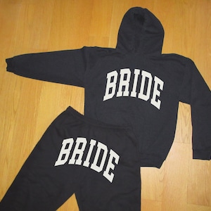 May include: A black hooded sweatshirt and matching sweatpants set. The word "BRIDE" is printed in large, white, block letters across the front of the sweatshirt and the sweatpants. The set is displayed on a wooden surface.