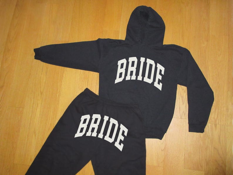 May include: A black hooded sweatshirt and matching sweatpants set. The word "BRIDE" is printed in large, white, block letters across the front of the sweatshirt and the sweatpants. The set is displayed on a wooden surface.