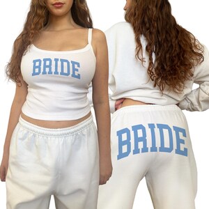 Pu&ograve; includere: Completo da salotto da sposa bianco con canotta e pantaloni della tuta. La canotta ha spalline sottili e la scritta "BRIDE" in lettere a blocchi azzurro chiaro. Anche i pantaloni della tuta mostrano "BRIDE" nello stesso carattere azzurro chiaro.