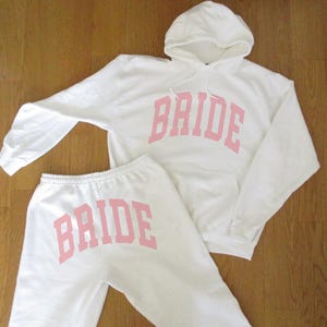 May include: A white hoodie and sweatpants set. The word "BRIDE" is printed in pink block letters across the chest of the hoodie and the seat of the sweatpants. The hoodie has a hood and long sleeves. The sweatpants have an elastic waistband.